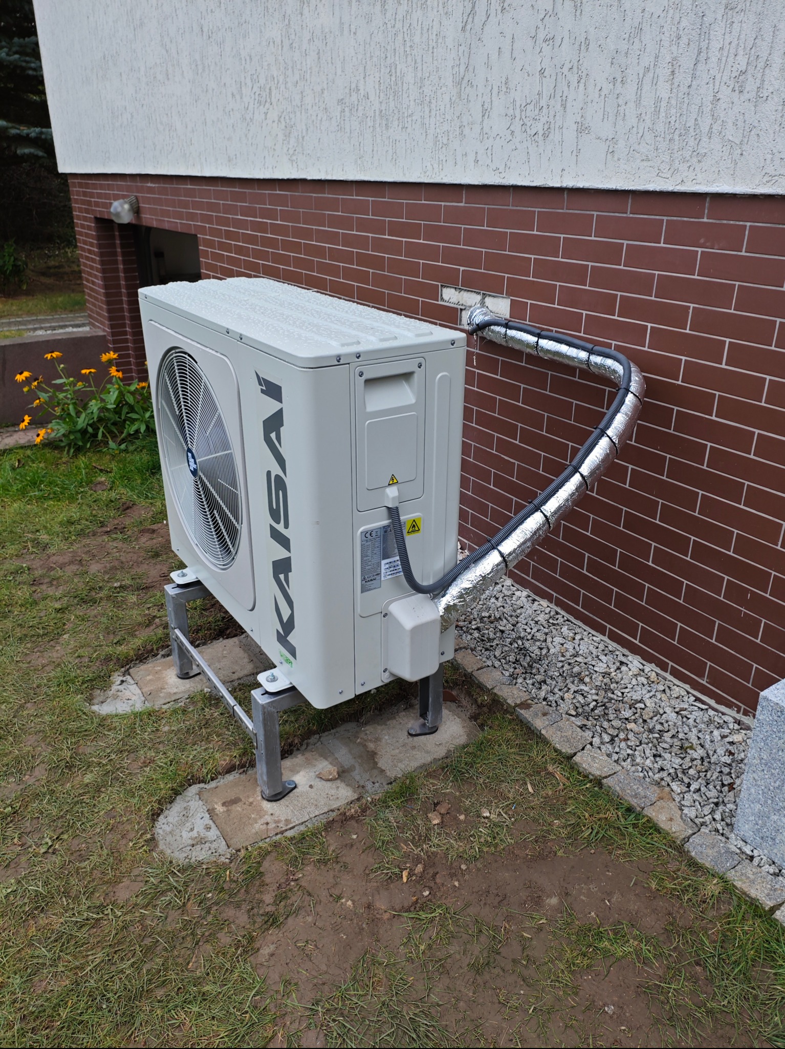 Zewnętrzna jednostka klimatyzacji Kaisai zamontowana na metalowym stelażu przy ścianie budynku z cegły, z izolowanymi rurami odprowadzającymi ciepło.