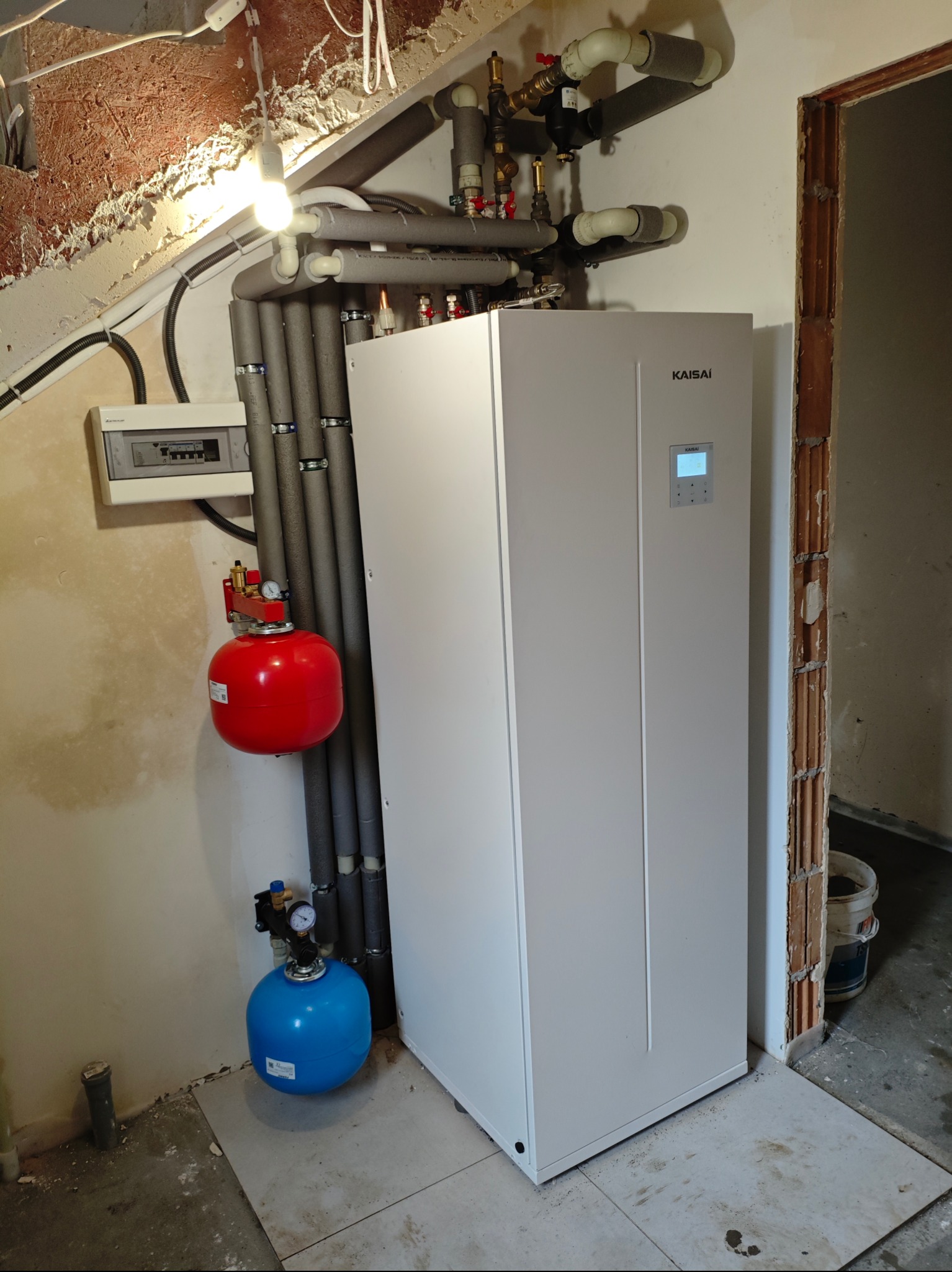 Biała pompa ciepła Kaisai z zintegrowanym zasobnikiem ciepłej wody użytkowej, widoczne rury instalacyjne w szarej otulinie, czerwony i niebieski zbiornik wyrównawczy, oraz skrzynka elektryczna...