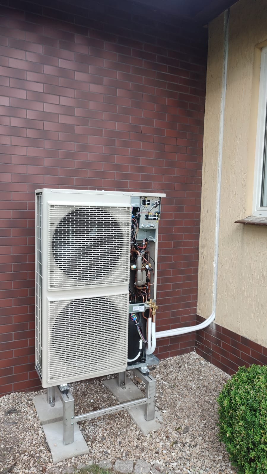 Zewnętrzna jednostka klimatyzacji dwu-wentylatorowa na metalowym stelażu, z otwartą obudową, eksponująca wewnętrzne komponenty, zamontowana przy ceglanym murze budynku.