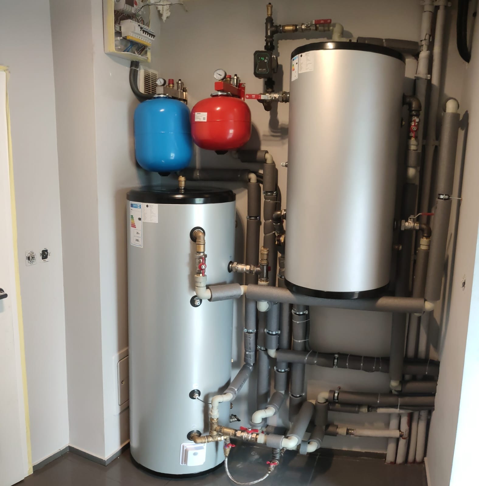 Dwa srebrne bojlery z niebieskim i czerwonym zbiornikiem wyrównawczym, połączone rurami z izolacją termiczną w kotłowni z szarą ścianą i podłogą.