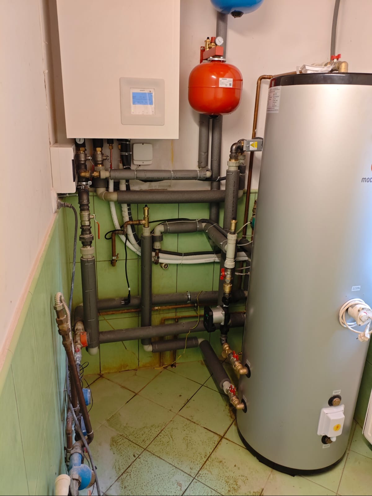 Kompletna instalacja centralnego ogrzewania z piecem gazowym, zasobnikiem ciepłej wody i rozbudowaną siecią rur w pomieszczeniu z zielonymi płytkami.