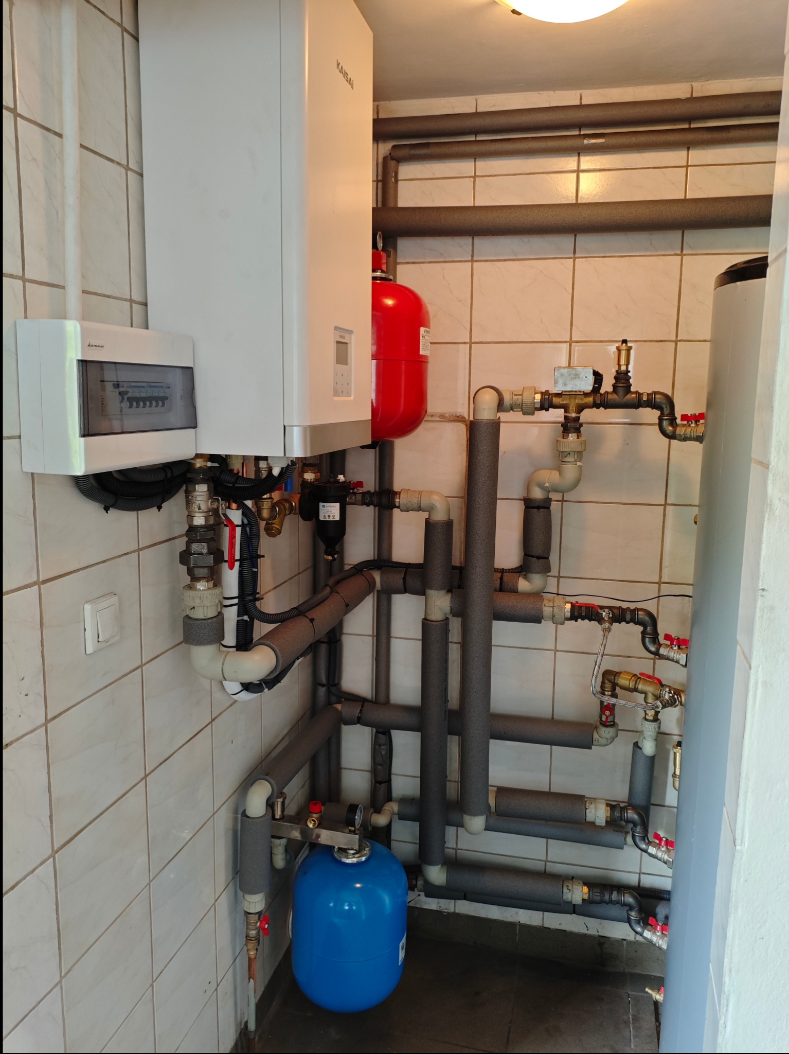 Węzeł cieplny w kotłowni z białym piecem gazowym Kaisai, czerwonym zbiornikiem wyrównawczym i niebieskim naczyniem przeponowym, liczne rury w izolacji termicznej, białe kafelki na ścianach.