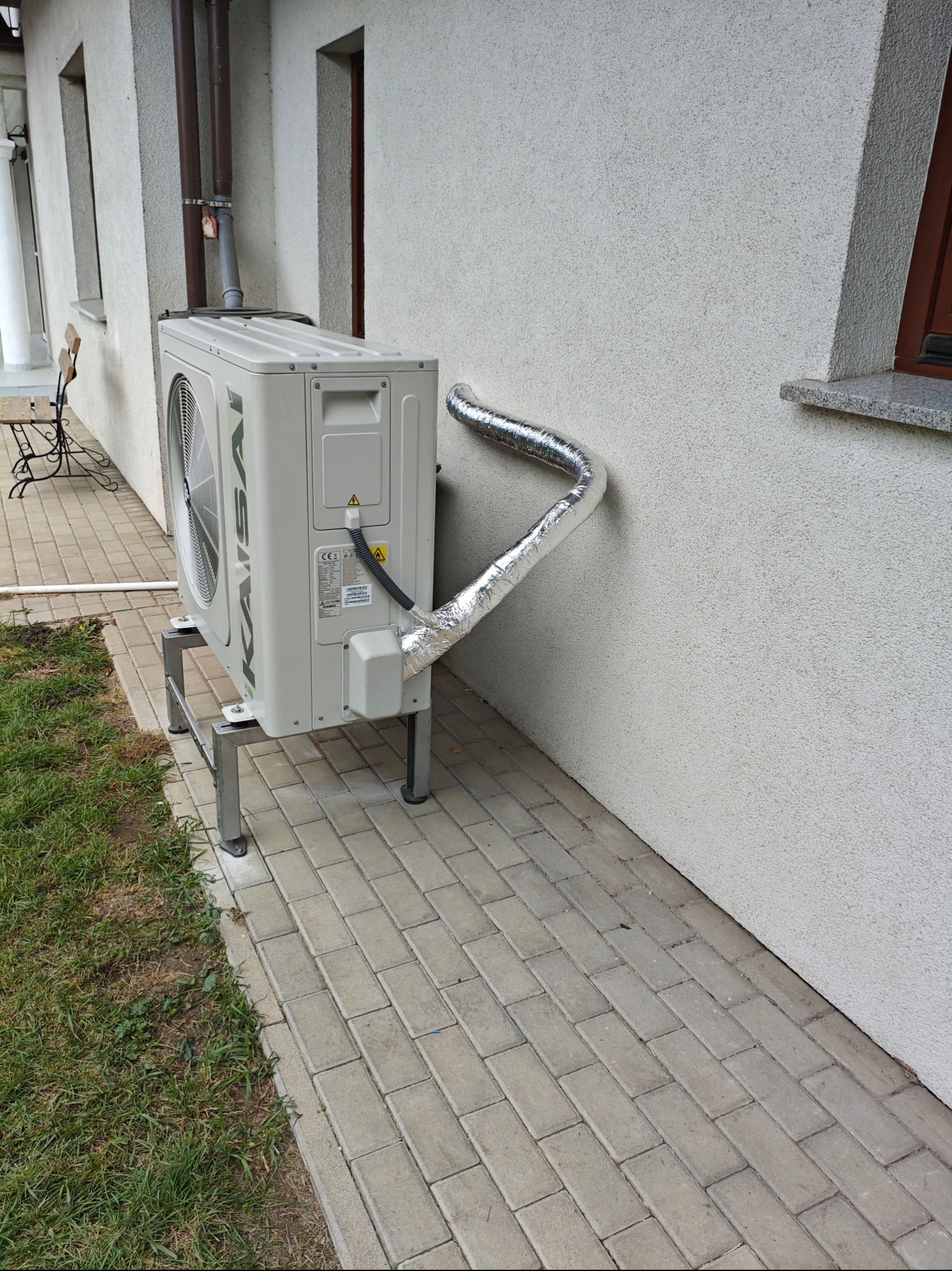 Zewnętrzna jednostka klimatyzacji zamontowana na metalowym stelażu przy ścianie budynku, z widocznym podłączeniem rury w srebrnej izolacji.