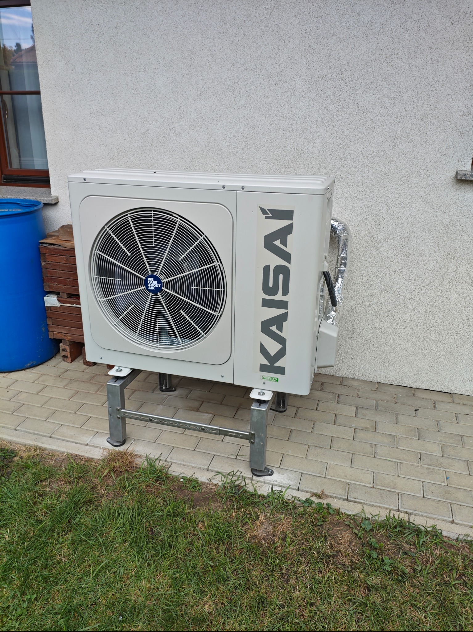Zewnętrzna jednostka klimatyzacji Kaisai zamontowana na metalowym stelażu przy ścianie budynku z widoczną trawą i kostką brukową.