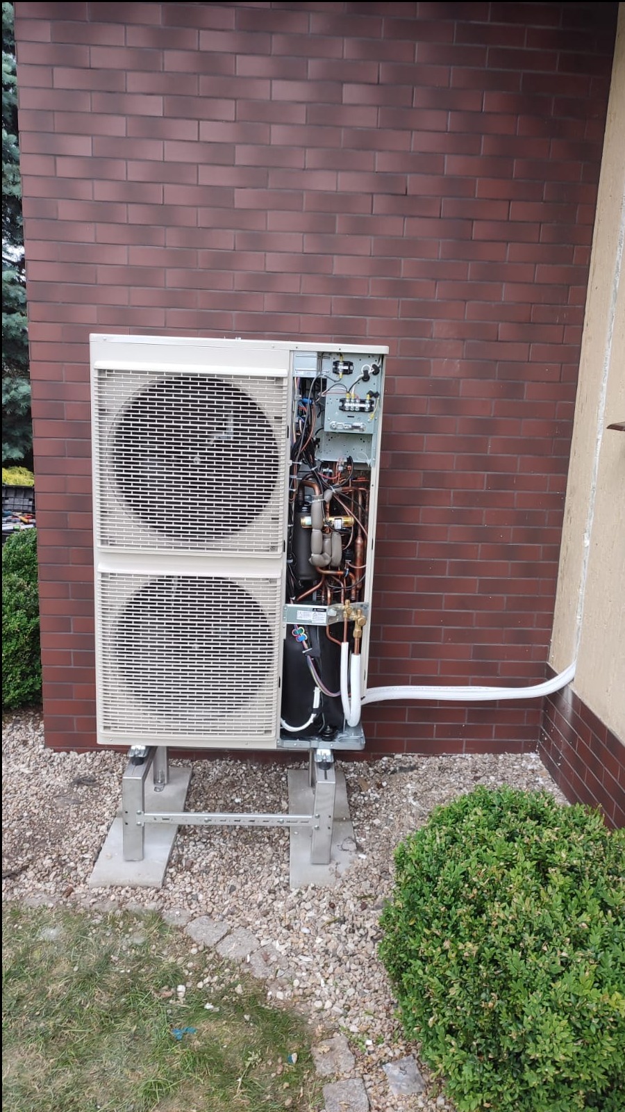 Zewnętrzna jednostka klimatyzacji typu split, z otwartą obudową ukazującą wewnętrzne komponenty, zamontowana na betonowych podstawach przed ceglaną ścianą budynku, z widocznymi przewodami...