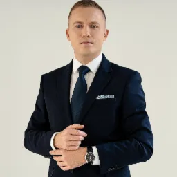 Portret mężczyzny w granatowym garniturze, białej koszuli i ciemnoniebieskim krawacie z wzorzystą poszetką, spoglądającego wprost, z zegarkiem na lewym nadgarstku.