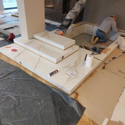 New Home, Kompleksowe Wykończenia Wnętrz - Prace wykończeniowe w trakcie: montaż płytek marmuropodobnych na ścianie i obudowie kominka, widoczne narzędzia i materiały budowlane, zabezpieczone schody folią.