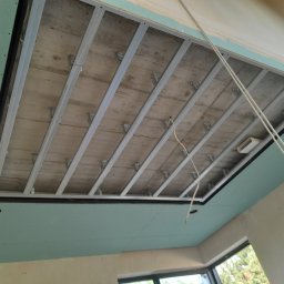 New Home, Kompleksowe Wykończenia Wnętrz - Sufit podwieszany z płyt kartonowo-gipsowych w trakcie montażu, widoczna metalowa konstrukcja nośna i wstępnie zamontowane płyty w kolorze jasnej zieleni z widocznymi otworami na oświetlenie.