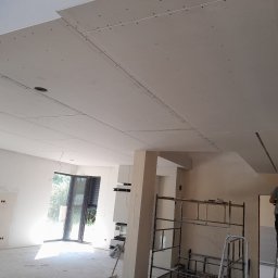 New Home, Kompleksowe Wykończenia Wnętrz - Montaż płyt kartonowo-gipsowych na suficie w przestronnym, jasnym pomieszczeniu; widoczny pracownik na drabinie, metalowe rusztowanie, surowe ściany i duże okno balkonowe.