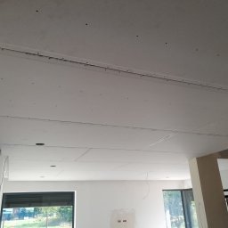 New Home, Kompleksowe Wykończenia Wnętrz - Sufit z zamontowaną płytą kartonowo-gipsową, widoczne łączenia płyt i wkręty, surowe wnętrze z oknami i elementami konstrukcyjnymi.