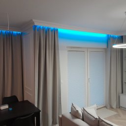 New Home, Kompleksowe Wykończenia Wnętrz - Nowoczesne wnętrze salonu z zasłonami, oświetleniem LED w listwach sufitowych, okrągłą lampą wiszącą i minimalistyczną zabudową meblową.