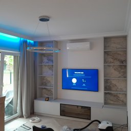 New Home, Kompleksowe Wykończenia Wnętrz - Nowoczesny salon z podświetleniem LED nad oknem, zabudową meblową z półkami wykończonymi marmurkowym wzorem, telewizorem na ścianie, oraz widocznym odkurzaczem i kartonem po sprzęcie.