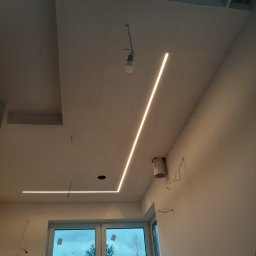 New Home, Kompleksowe Wykończenia Wnętrz - Nowoczesne oświetlenie LED w kształcie litery L na suficie podwieszanym w trakcie montażu, widoczne przewody i puszka elektryczna, okno z widokiem na drzewa.