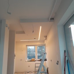 New Home, Kompleksowe Wykończenia Wnętrz - Wnętrze w trakcie prac wykończeniowych: białe ściany, sufit z oświetleniem LED, drabina aluminiowa, widoczne kable i przygotowania do montażu oświetlenia.
