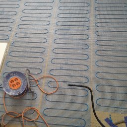 New Home, Kompleksowe Wykończenia Wnętrz - Rozłożona mata grzewcza na podłodze z widocznym przewodem zasilającym i przedłużaczem bębnowym.