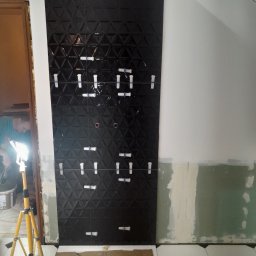 New Home, Kompleksowe Wykończenia Wnętrz - Ściana z czarnych, trójwymiarowych płytek, częściowo ułożonych z użyciem klipsów poziomujących, widoczny pracownik i lampa budowlana.