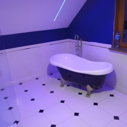 New Home, Kompleksowe Wykończenia Wnętrz - Nowoczesna łazienka na poddaszu z wolnostojącą wanną na złotych nóżkach, biało-czarna podłoga w kratkę, niebieska ściana i oświetlenie LED.