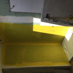 New Home, Kompleksowe Wykończenia Wnętrz - Wnętrze w trakcie prac: fragment ściany pomalowanej na żółto, częściowo wykończona ściana z płytkami, poziomica i fragment drewnianej podłogi, widoczne instalacje elektryczne.