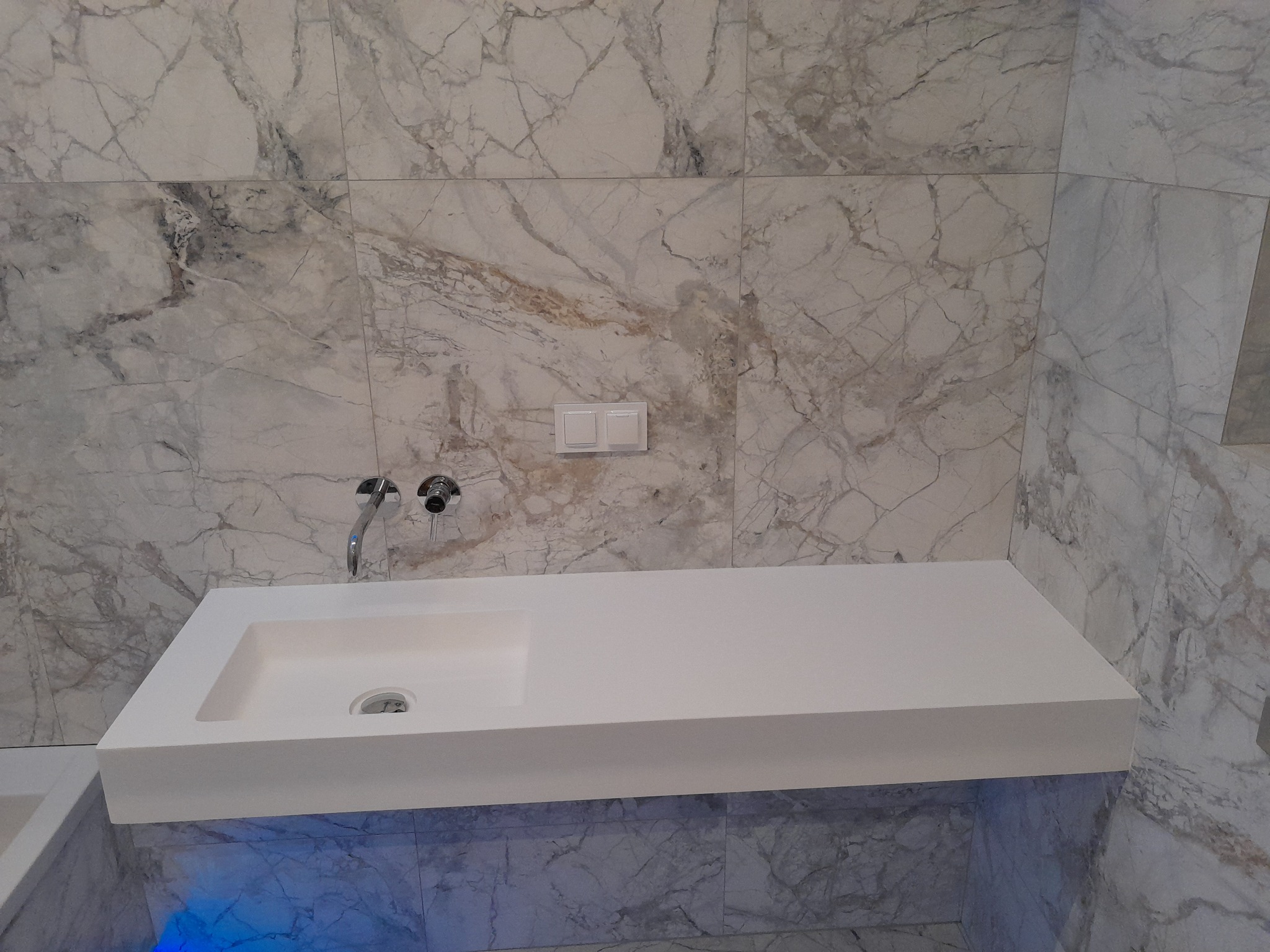 Nowoczesna łazienka z umywalką wpuszczaną w blat, podświetleniem LED i marmurowymi płytkami na ścianach.