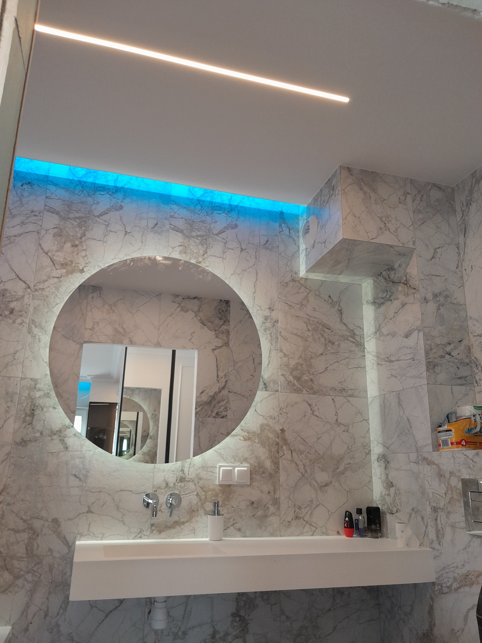 Nowoczesna łazienka z marmurowymi płytkami, okrągłym lustrem z podświetleniem LED i minimalistyczną umywalką.