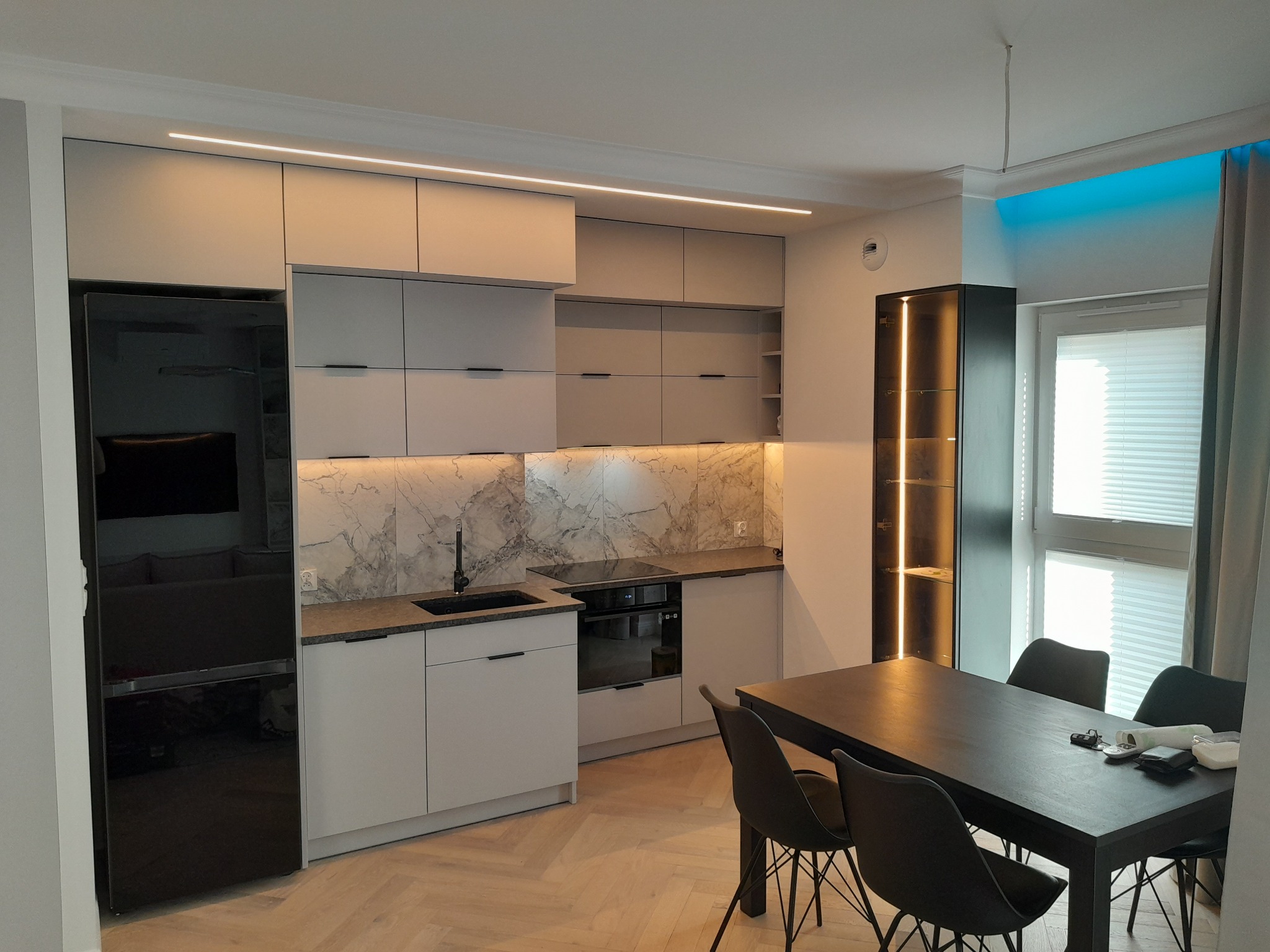 Nowoczesna kuchnia z szarymi szafkami, marmurowym backsplashiem i czarnym sprzętem AGD, oświetlona listwami LED, z widocznym stołem jadalnym z czarnymi krzesłami.