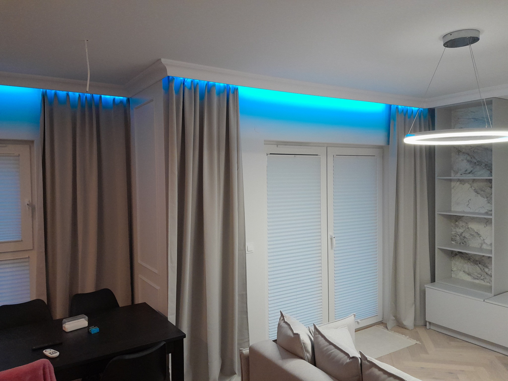 Nowoczesne wnętrze salonu z zasłonami, oświetleniem LED w listwach sufitowych, okrągłą lampą wiszącą i minimalistyczną zabudową meblową.