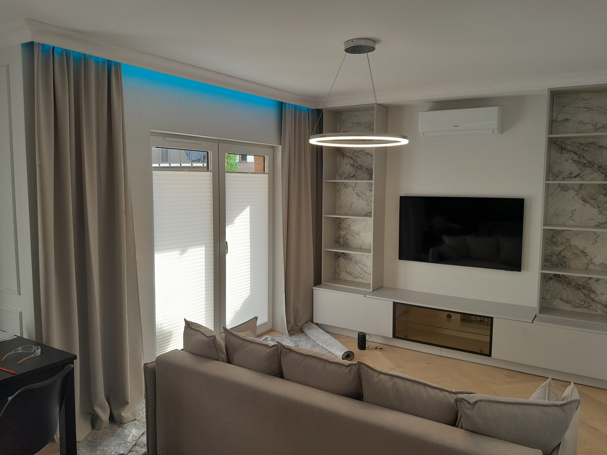 Jasne wnętrze salonu w Warszawie z szarym narożnikiem, podświetleniem LED nad oknem, zabudową meblową z imitacją marmuru i okrągłą lampą wiszącą.