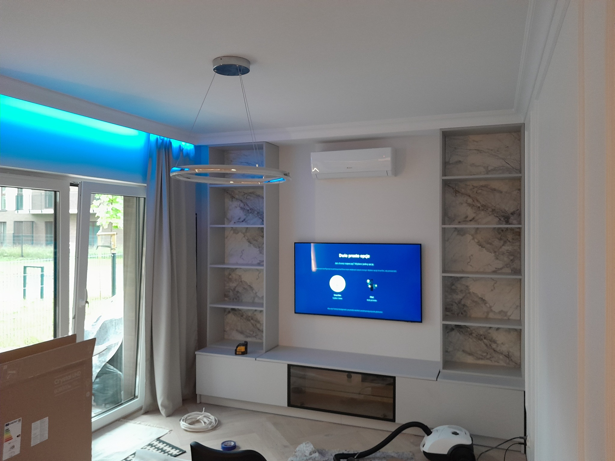 Nowoczesny salon z podświetleniem LED nad oknem, zabudową meblową z półkami wykończonymi marmurkowym wzorem, telewizorem na ścianie, oraz widocznym odkurzaczem i kartonem po sprzęcie.