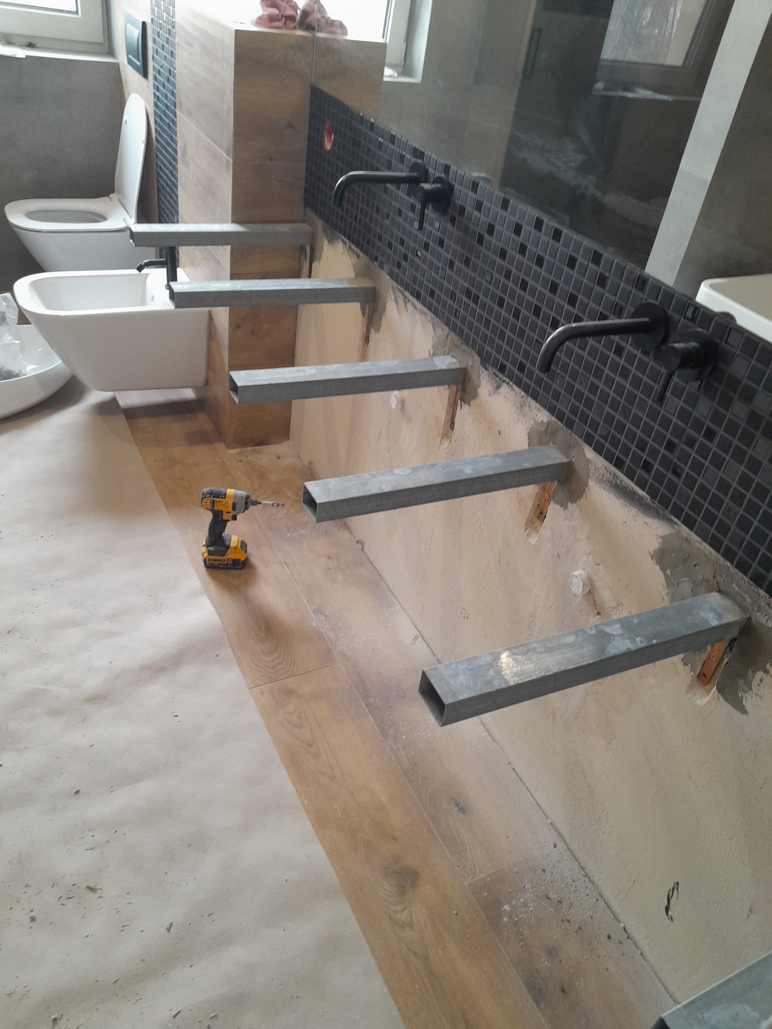 Łazienka w trakcie montażu umywalek: widoczne metalowe wsporniki przymocowane do ściany z mozaiką, bateria, toaleta i bidet. Na podłodze częściowo rozłożona folia ochronna, obok wiertarka.