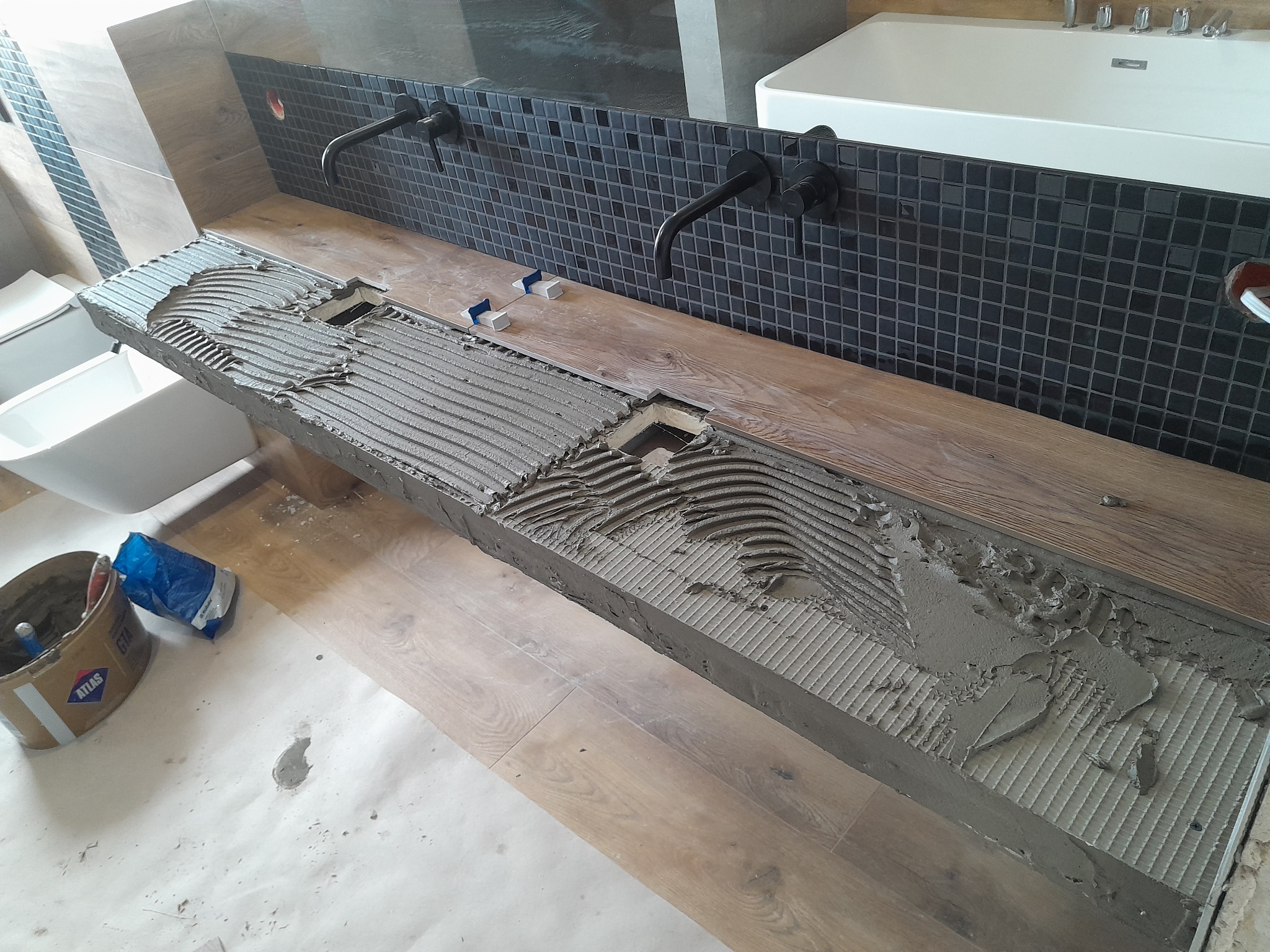 Montaż blatu z płytek imitujących drewno w łazience, widoczny klej do płytek, czarne baterie umywalkowe i mozaika na ścianie, w tle wanna.