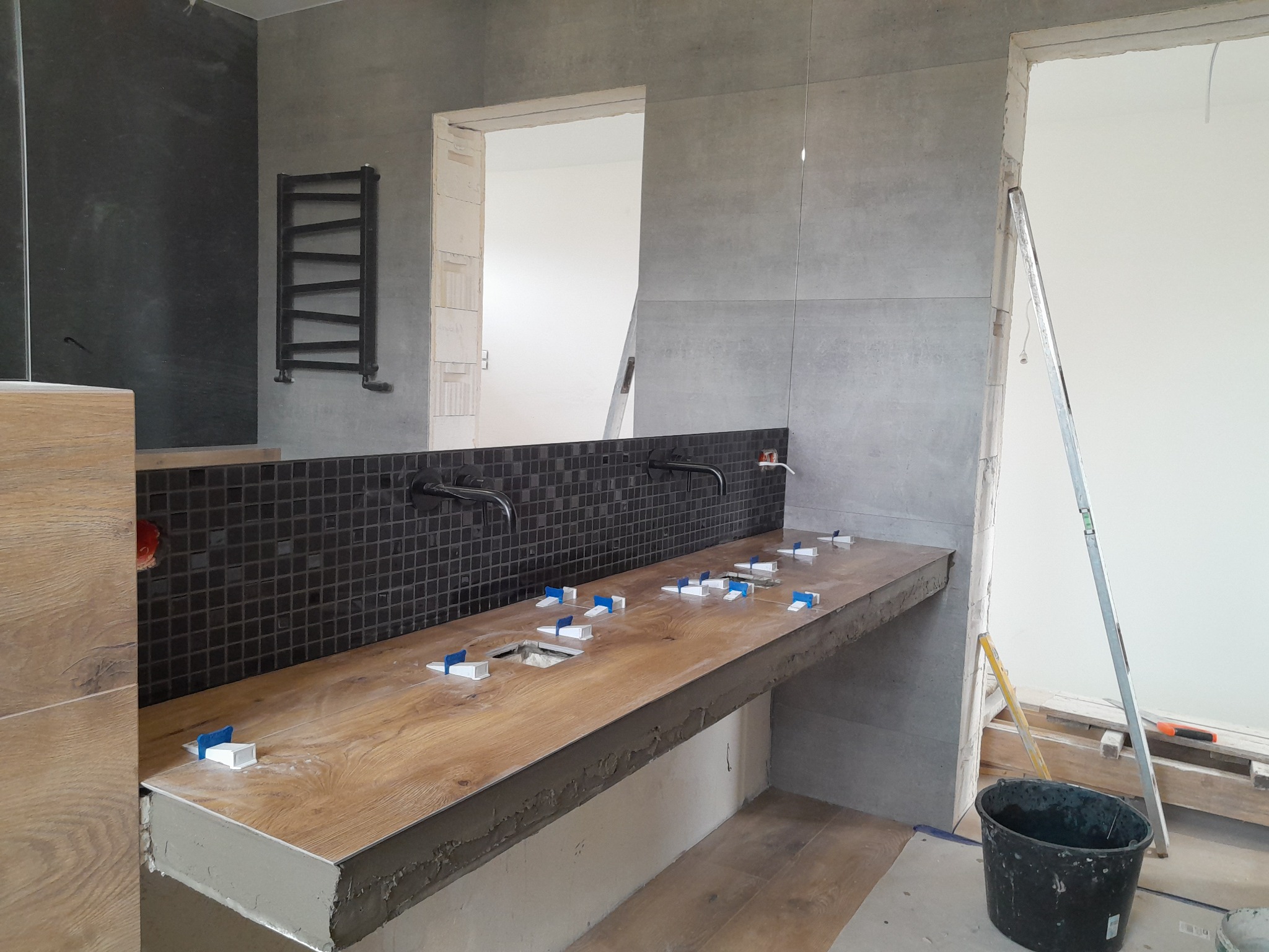 Łazienka w trakcie wykańczania: drewnopodobny blat pod umywalki z czarną mozaiką na ścianie, widoczne krzyżyki glazurnicze, surowy stan wykończenia ścian, grzejnik drabinkowy na ścianie.