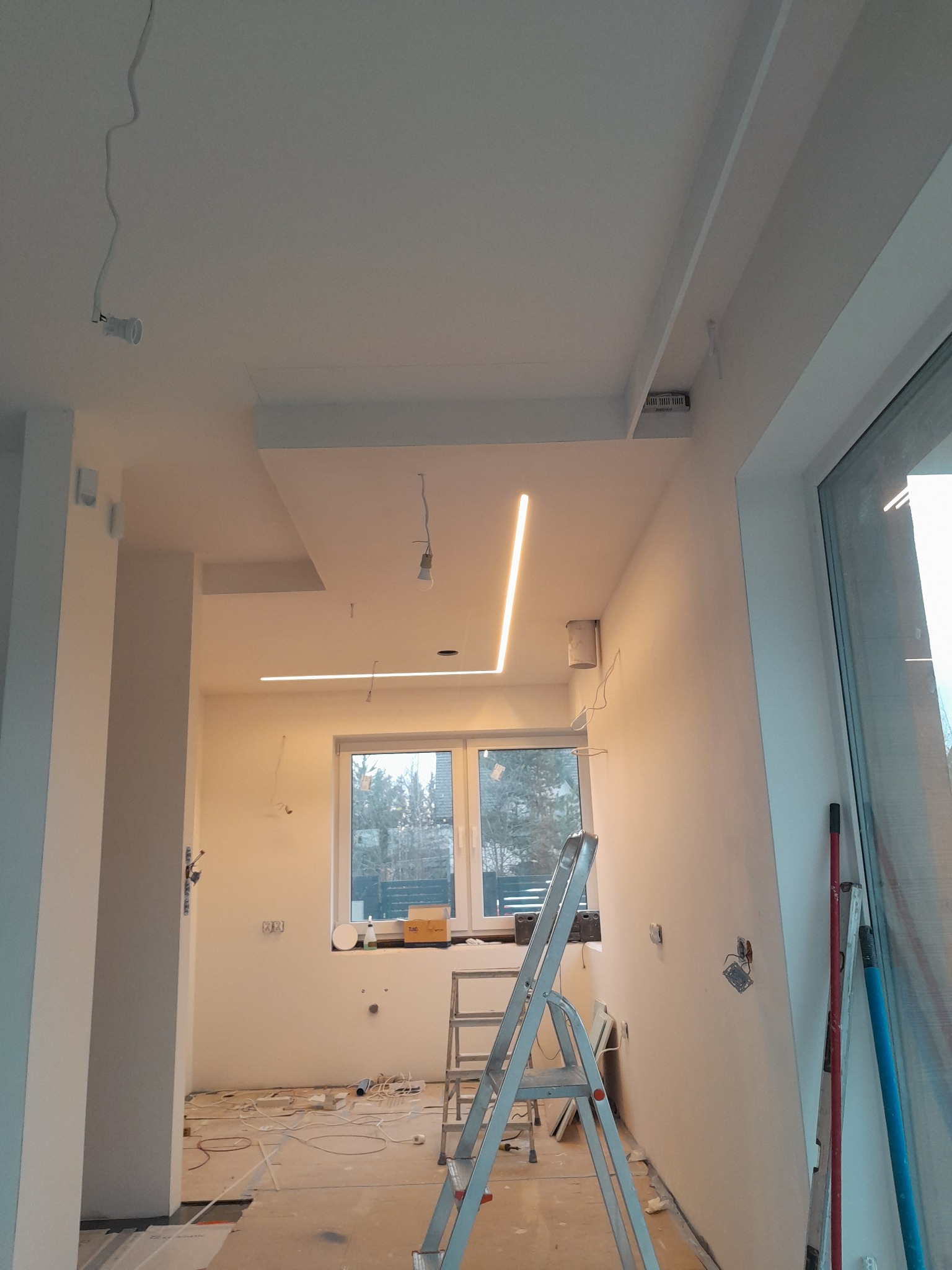 Wnętrze w trakcie prac wykończeniowych: białe ściany, sufit z oświetleniem LED, drabina aluminiowa, widoczne kable i przygotowania do montażu oświetlenia.