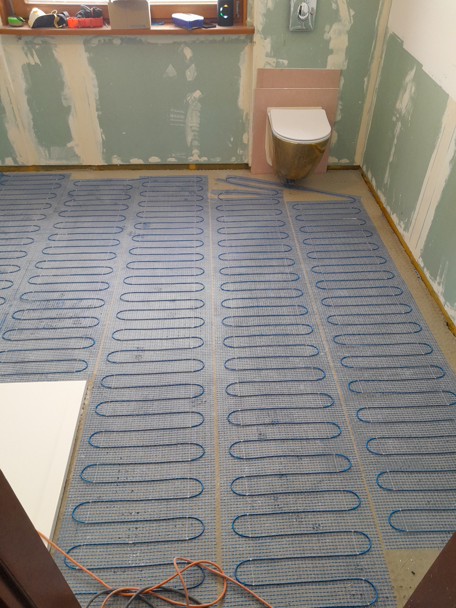 Nierówna podłoga łazienki z rozłożoną siatką grzewczą przed wylaniem wylewki, ściany w surowym stanie, częściowo pokryte płytami gipsowo-kartonowymi, wisząca toaleta w tle.