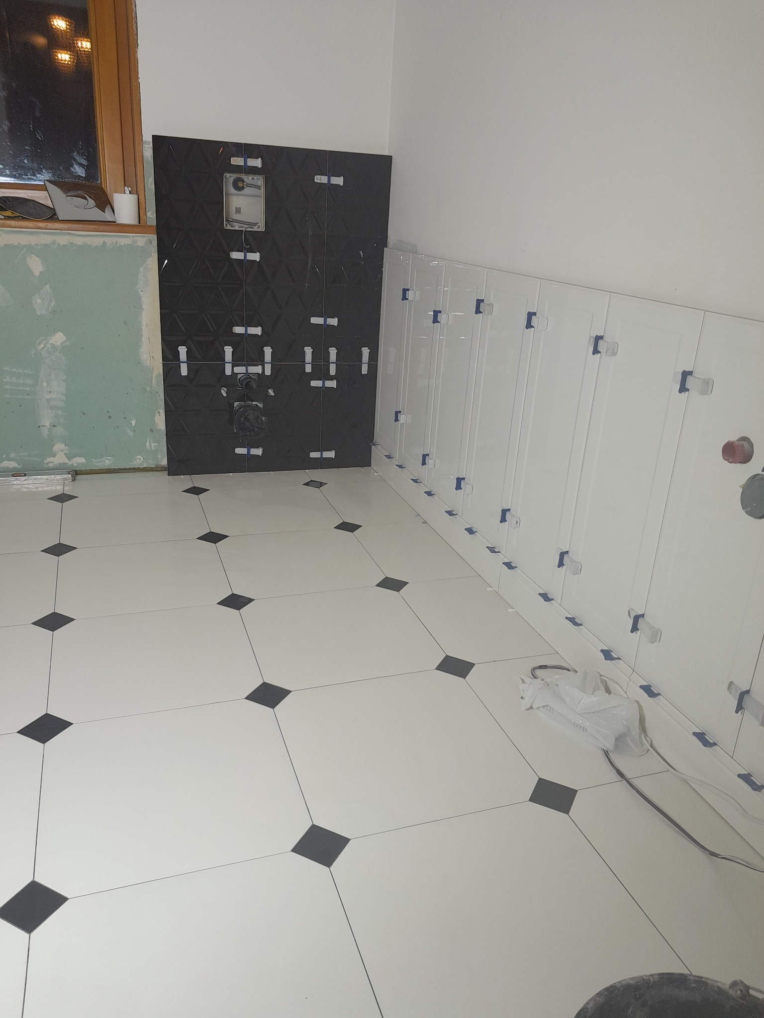 Łazienka w trakcie remontu: biało-czarna podłoga z płytek, czarne płytki na ścianie z zamontowaną armaturą, białe płytki na ścianie z widocznymi krzyżykami dystansowymi.