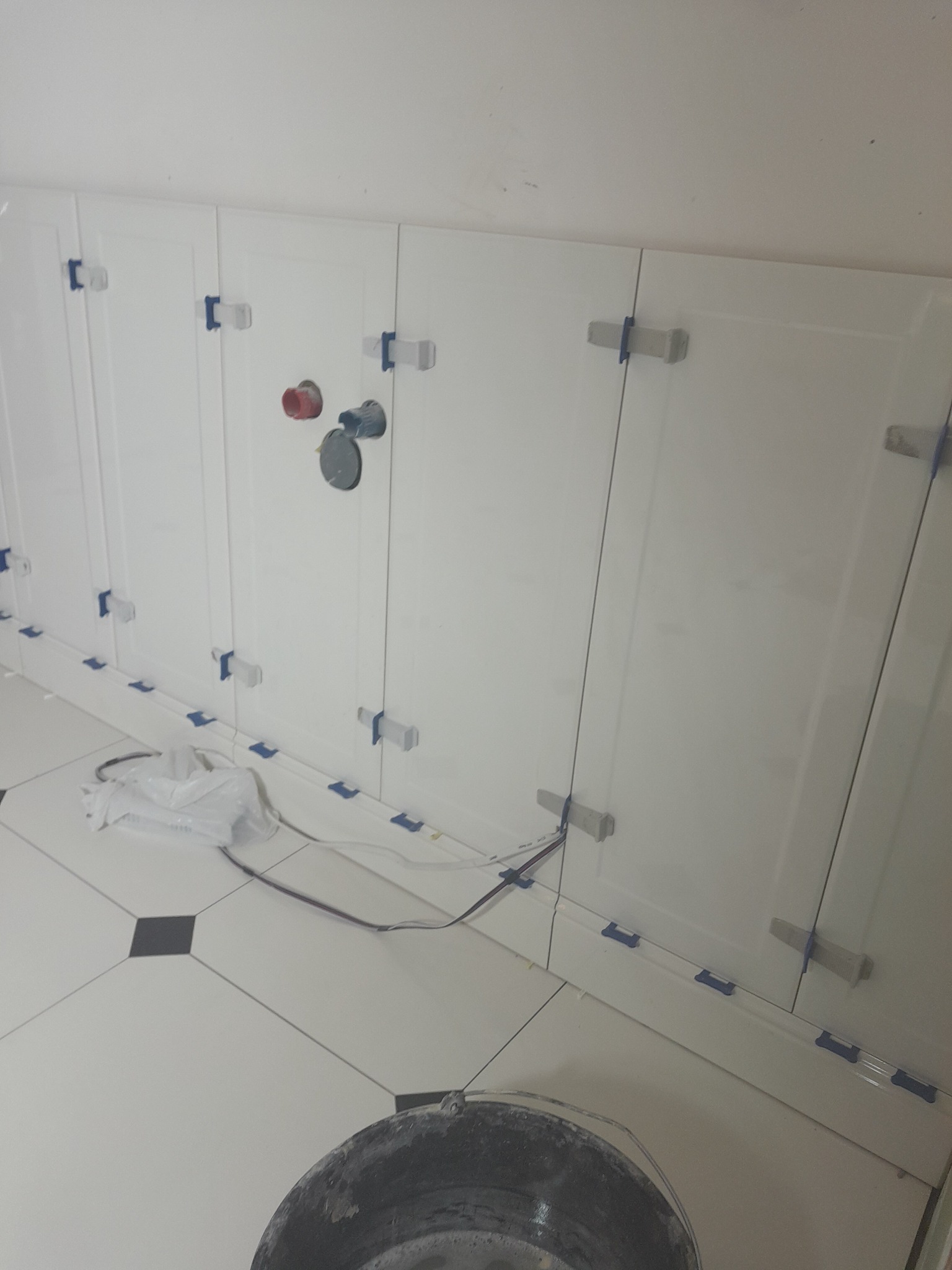 Białe płytki ścienne z elementami mocującymi i niebieską taśmą zabezpieczającą, widoczne rury i wiadro na podłodze z białych i czarnych płytek.