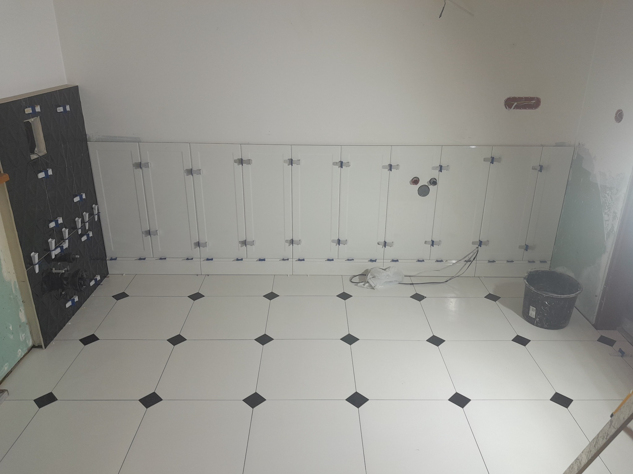 Łazienka w trakcie układania płytek: białe płytki podłogowe z czarnymi rombami, częściowo wykończona ściana z białymi płytkami i czarna ściana z elementami instalacji sanitarnych, widoczne krzyżyki...