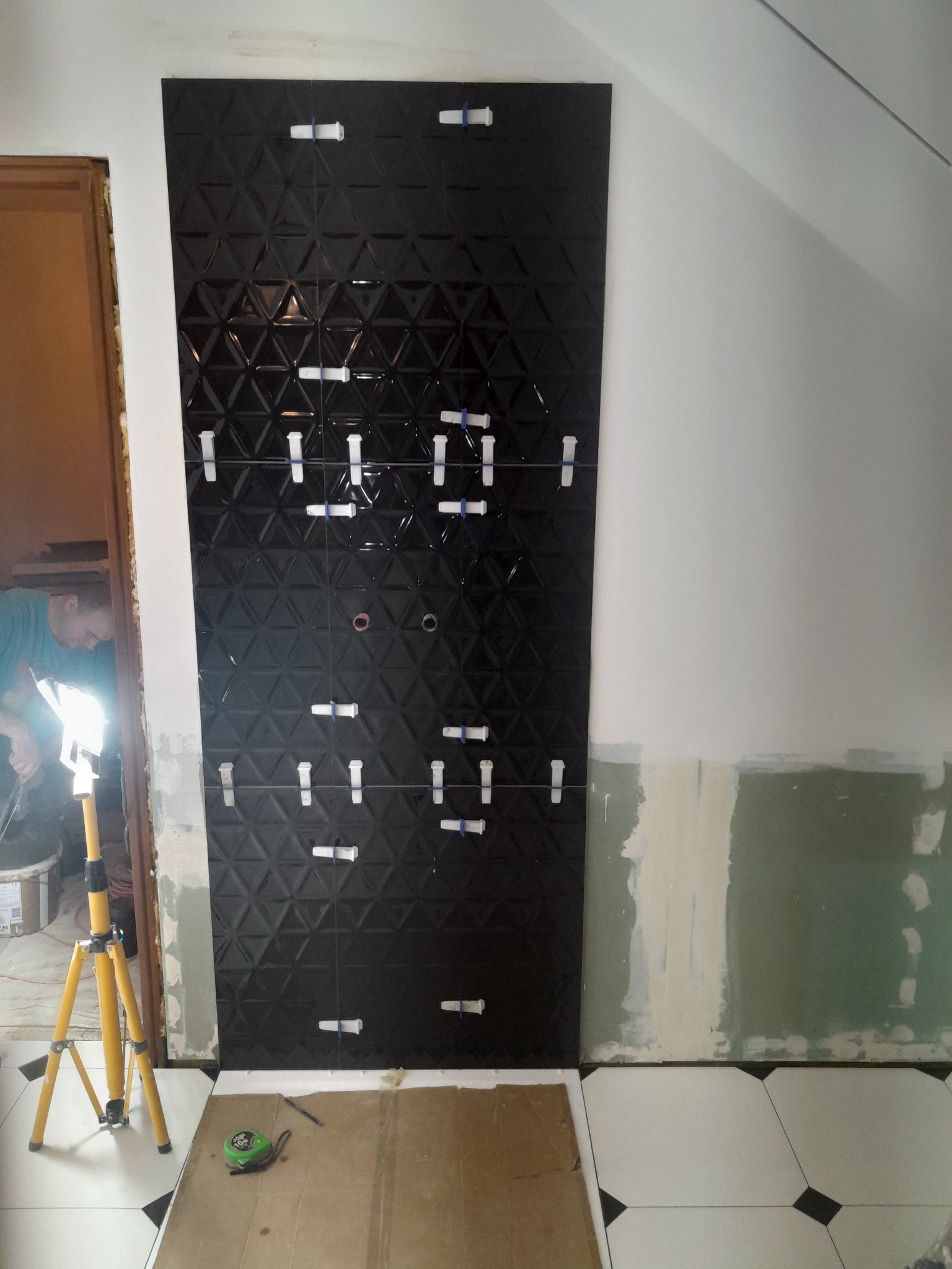 Ściana z czarnych, trójwymiarowych płytek, częściowo ułożonych z użyciem klipsów poziomujących, widoczny pracownik i lampa budowlana.