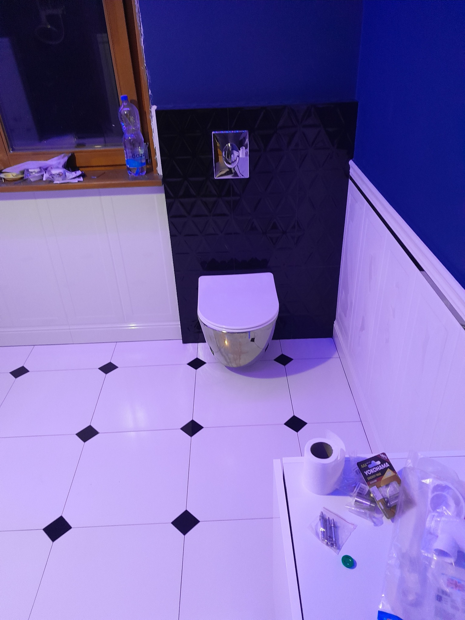 Nowoczesna łazienka w trakcie wykańczania: biało-czarne płytki podłogowe, czarna ściana z geometrycznym wzorem, częściowo zamontowana toaleta, akcesoria instalacyjne na blacie.