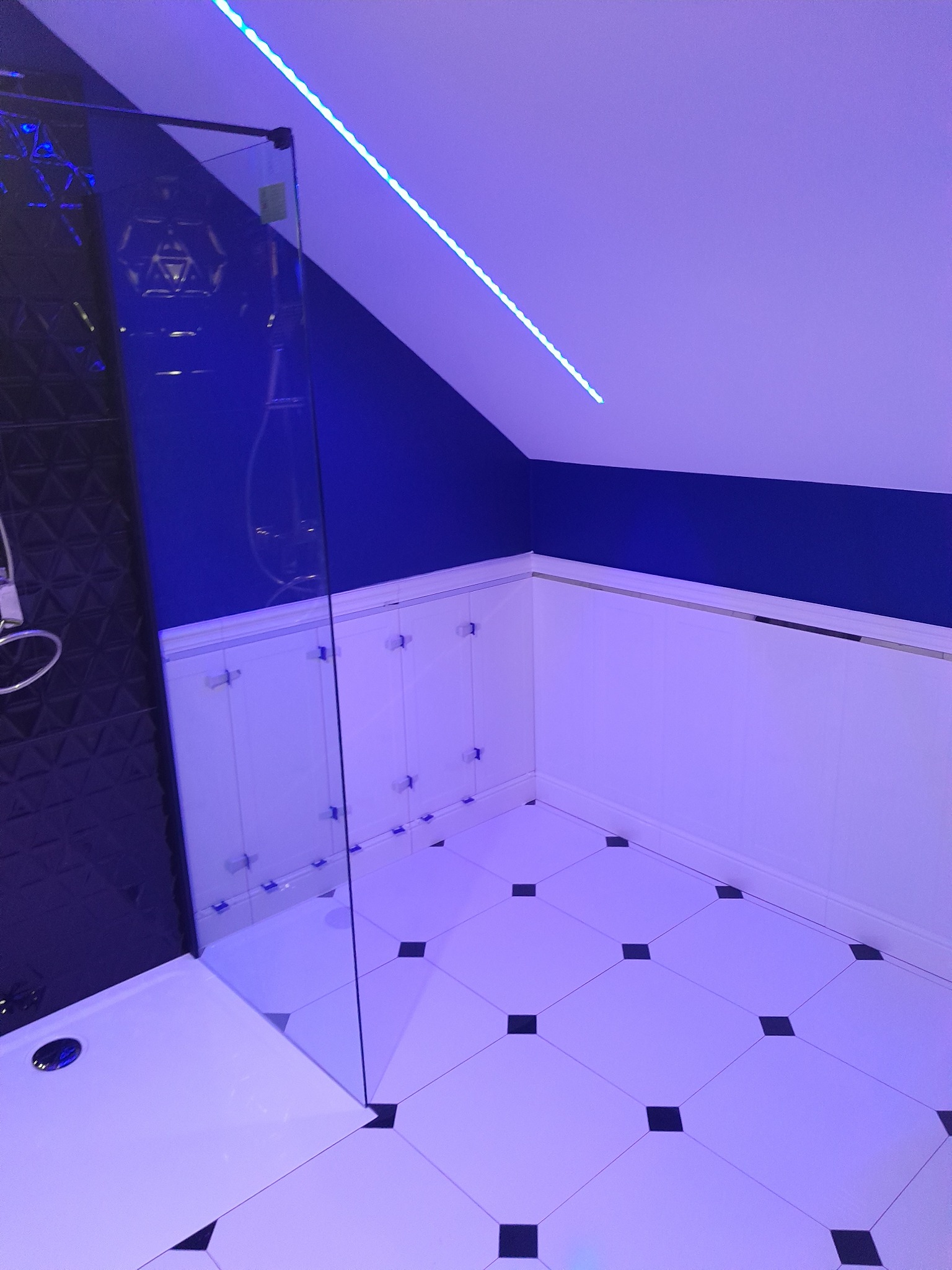 Nowoczesna łazienka z kabiną prysznicową ze szklaną ścianką, czarno-białymi płytkami podłogowymi i niebieskim oświetleniem LED podsufitowym.