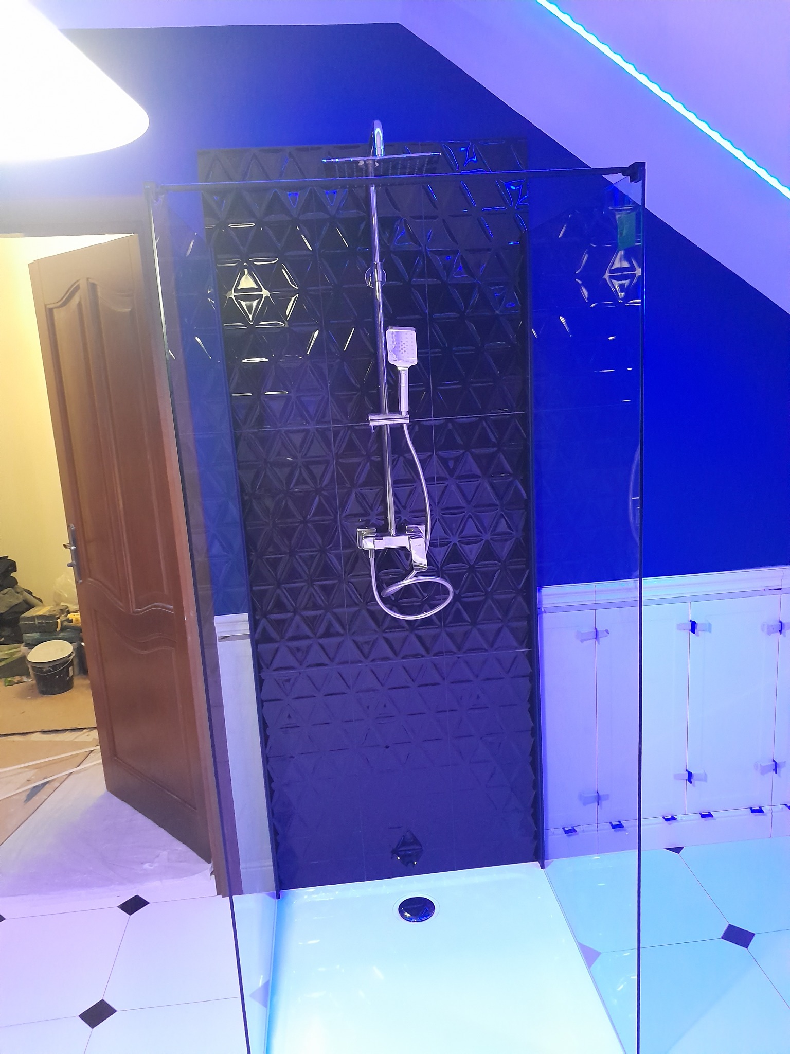 Nowoczesna kabina prysznicowa z czarnymi, trójwymiarowymi płytkami i chromowaną armaturą, oświetlona niebieskim światłem LED, widoczna częściowo otwarta drewniana rama drzwi i wiadro z farbą.