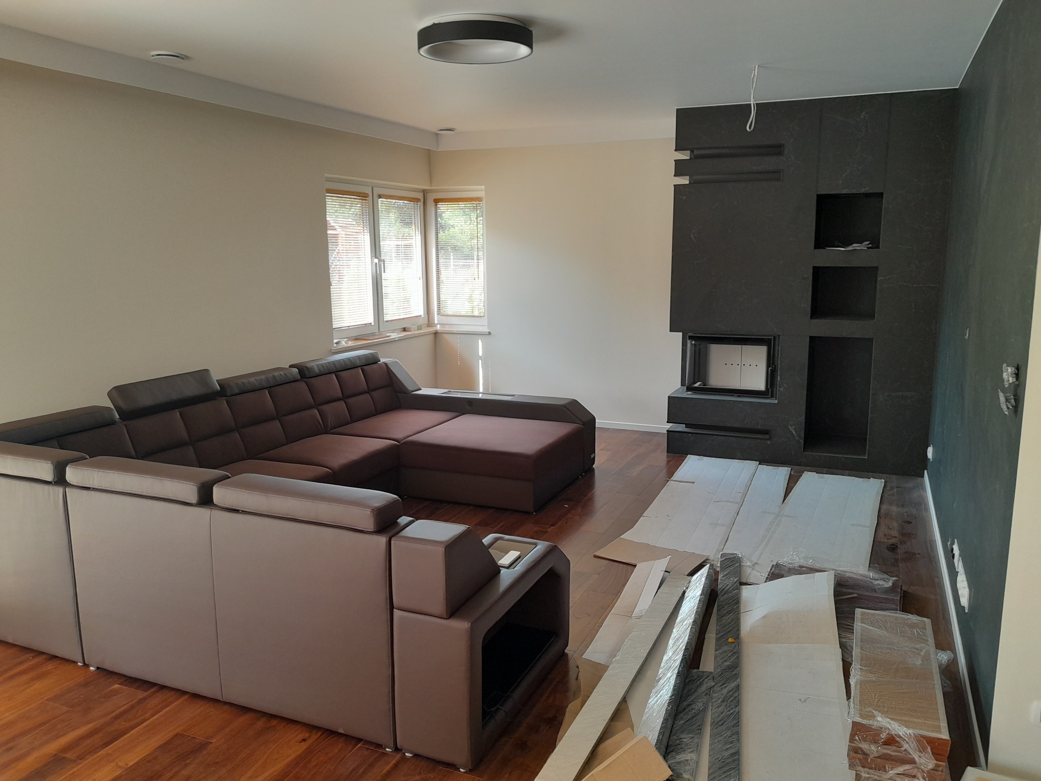 Salon w trakcie prac wykończeniowych: brązowy narożnik, okno z żaluzjami, ściana z kominkiem wykończona ciemnym kamieniem, zabezpieczona podłoga, widoczne listwy przypodłogowe.