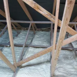 Poddasze z drewnianą konstrukcją pokryte warstwą jasnej pianki izolacyjnej, widoczne elementy mocujące konstrukcję.