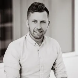 Portret uśmiechniętego mężczyzny w jasnej koszuli na tle okna, fotografia w odcieniach szarości z delikatnym ziarnem.