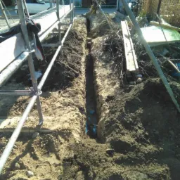 Wykop pod instalację w Nowym Dworze Gdańskim, widoczne niebieskie rury w ziemi, częściowo zasypane, rusztowanie w tle.