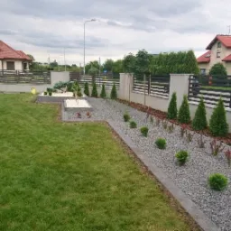 Nowoczesny ogród z równo przystrzyżonym trawnikiem, ścieżką z szarego żwiru obramowaną kostką, ozdobnymi krzewami i tujami wzdłuż ogrodzenia.