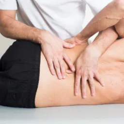 Mężczyzna leży na brzuchu na stole do masażu, a specjalista w Knurowie masuje mu plecy. Pacjent trzyma rękę na głowie.