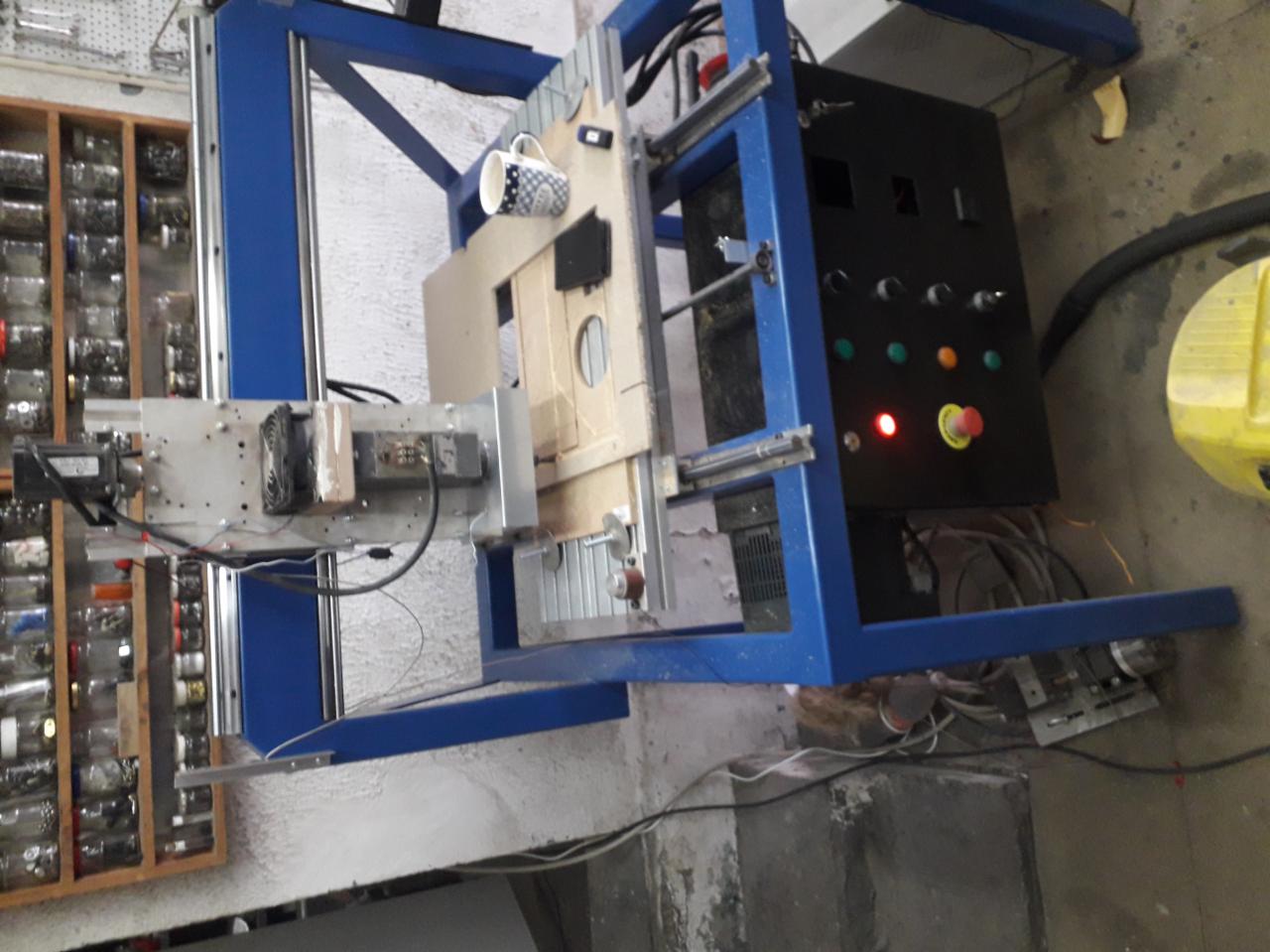 Niebieska maszyna CNC z kubkiem w kropki na ramie, panel sterowania z przyciskami i awaryjnym wyłącznikiem, obok regał z sortowanymi śrubkami i nakrętkami.