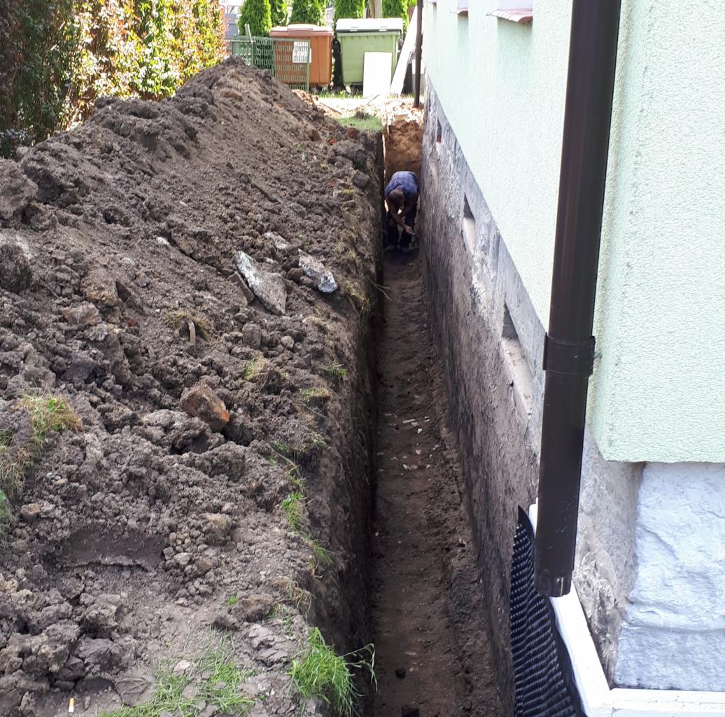 Wąski wykop wzdłuż fundamentu budynku, widoczna osoba kopiąca na dnie wykopu, zabezpieczenie fundamentu folią kubełkową.