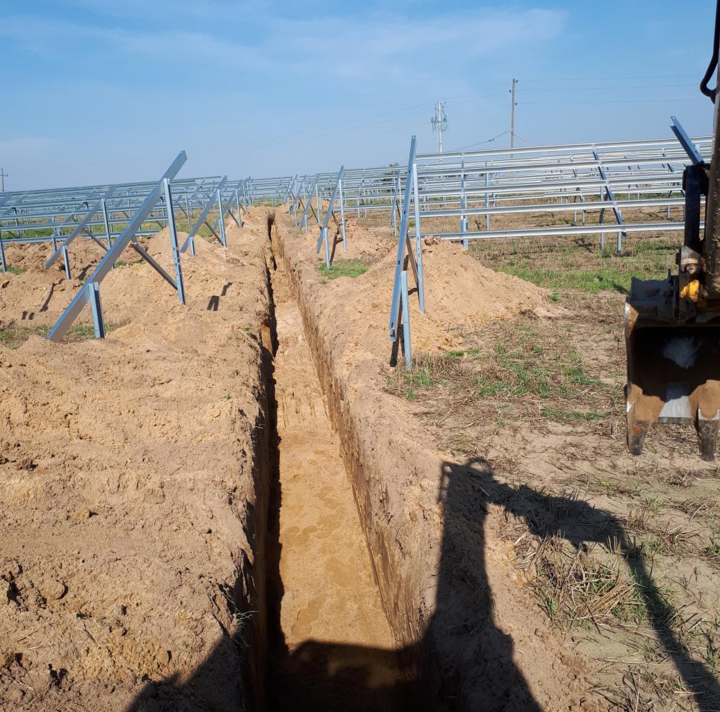 Wykopany wąski rów w piaszczystej glebie pod instalację, widoczne metalowe konstrukcje wsporcze, cień koparki na pierwszym planie, błękitne niebo.