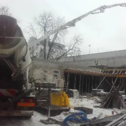 Betonomieszarka z pompą podaje beton na budowę w zimowy dzień, widoczni pracownicy w żółtych kamizelkach, szalunki przygotowane do wylewania betonu.